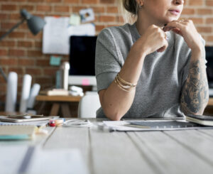 Young Millennial or Gen-Z professional woman with tattoos and a thoughtful expression, sitting in a modern office and working independently